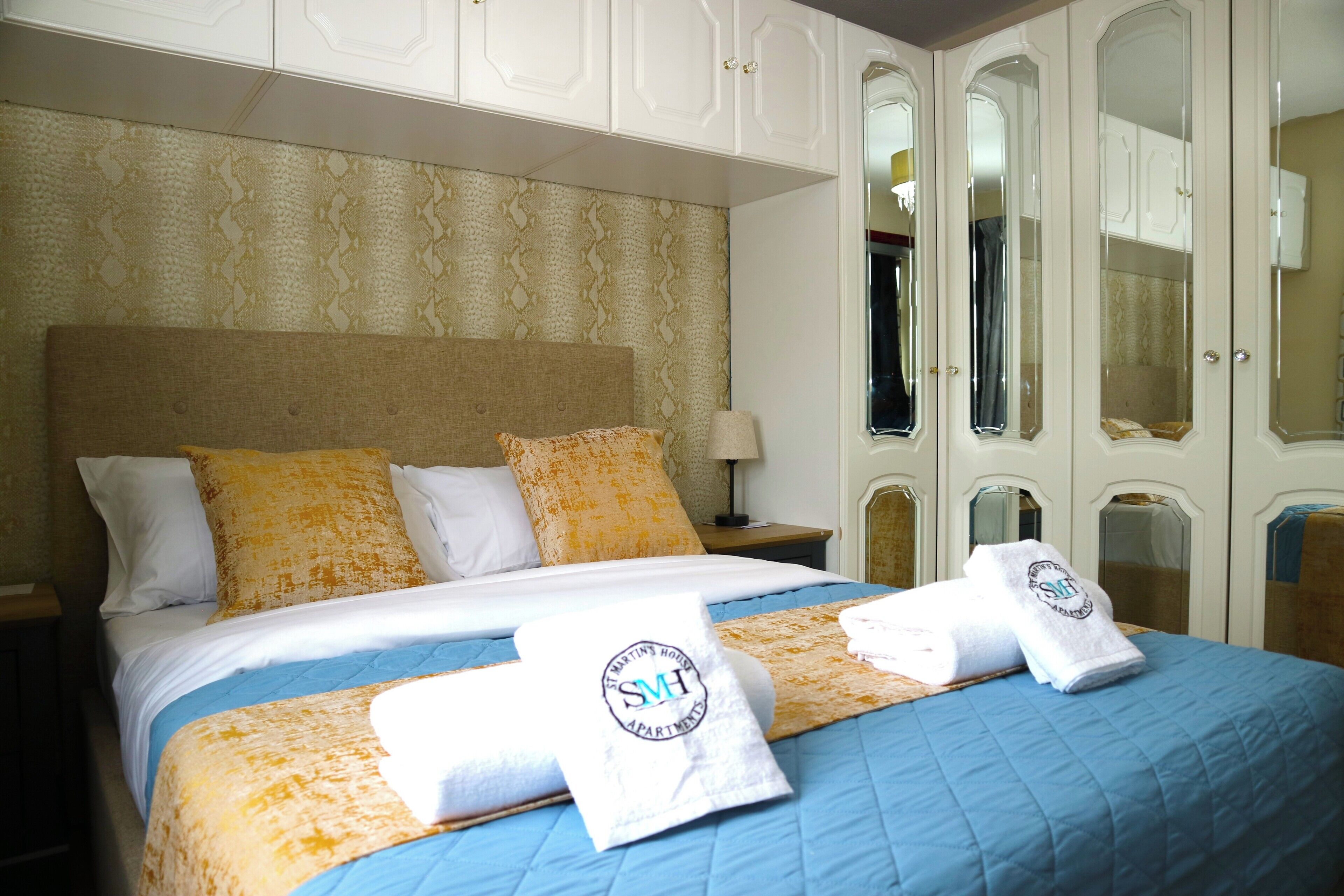 Une chambre d’hôtel avec un grand lit, une literie blanche et jaune à motifs, une table de chevet avec une lampe et une armoire aux portes miroir.