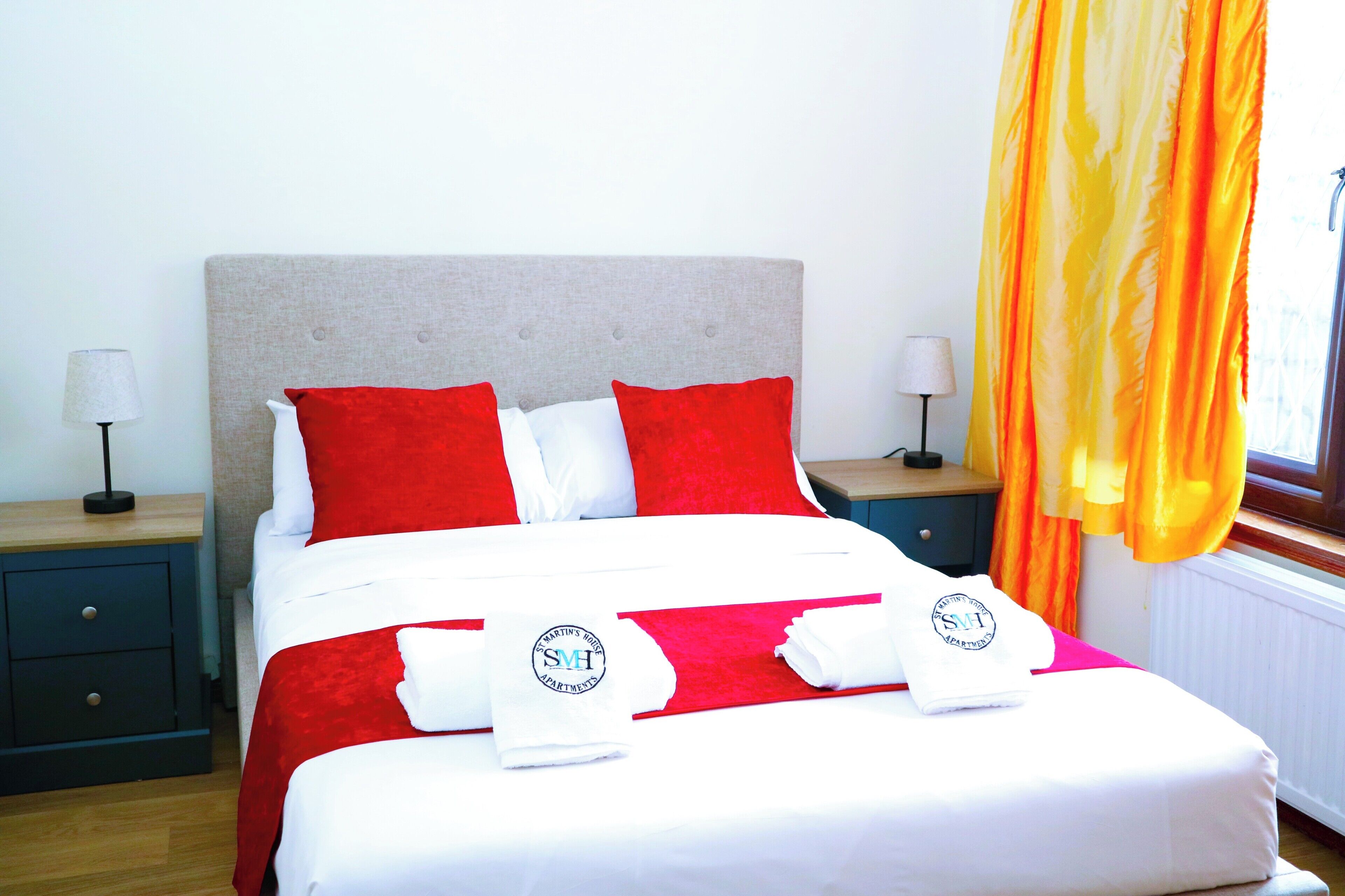 A neatly made bed with red and white bedding, two pillows, and towels with a logo. There are two bedside tables with lamps, a window with curtains, and a radiator.