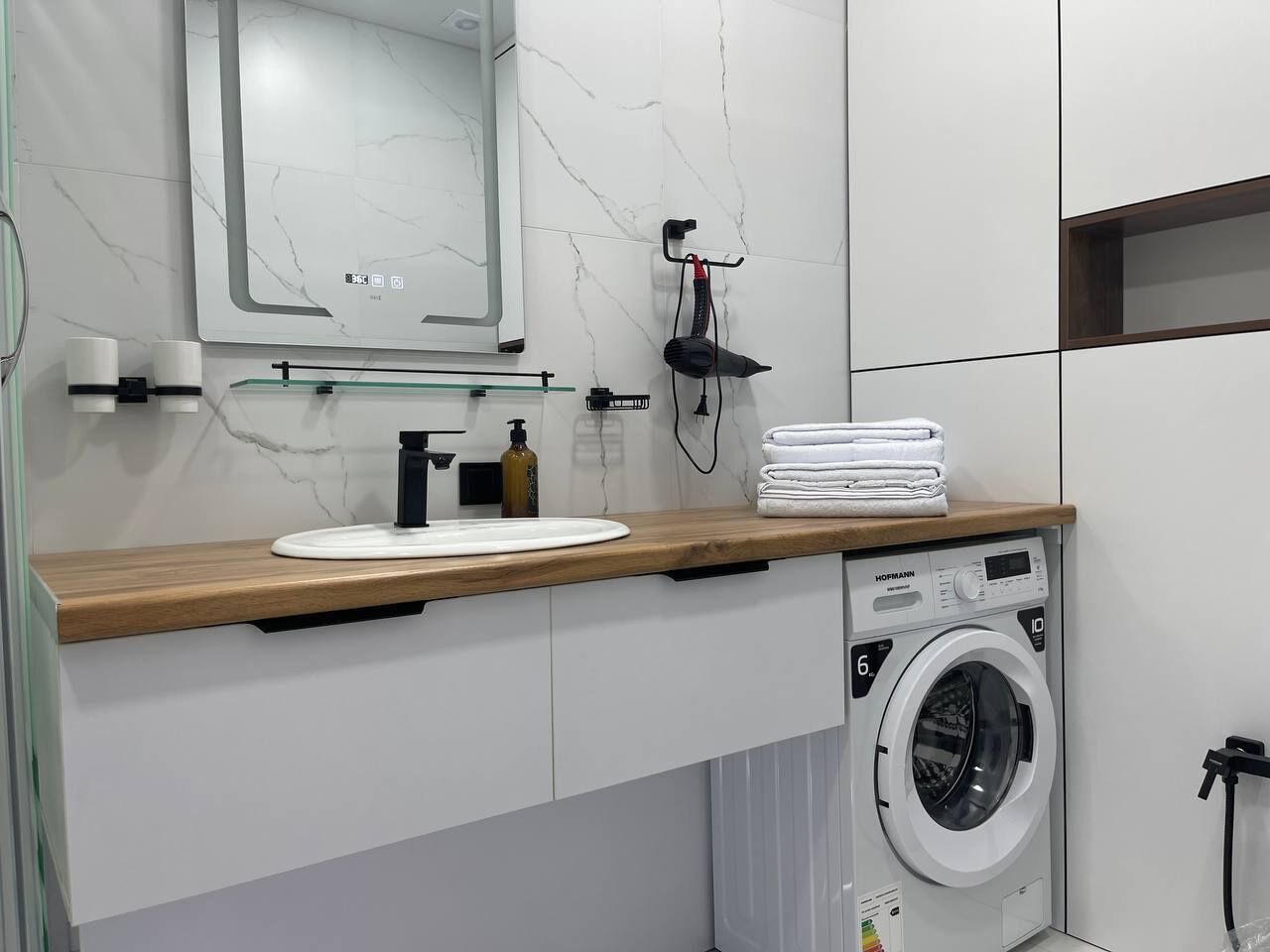 Une salle de bain moderne avec un pare-feu en marbre, un plan de travail en bois, un lavabo, un miroir, un sèche-cheveux et un lave-linge.