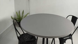 Une table ronde entourée de quatre chaises noires, un pot de fleurs et deux étagères murales ornées de petites plantes et d’un haut-parleur.