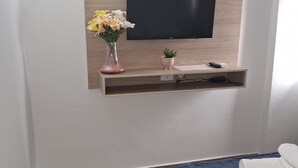 Une chambre avec un lit, un téléviseur fixé au mur, une petite étagère avec un vase de fleurs et une télécommande.