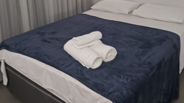 Un lit bien fait, avec une couvre-lit bleue et des oreillers blancs, une pile de serviettes pliées posée sur le lit, et une fenêtre dont les rideaux sont tirés.