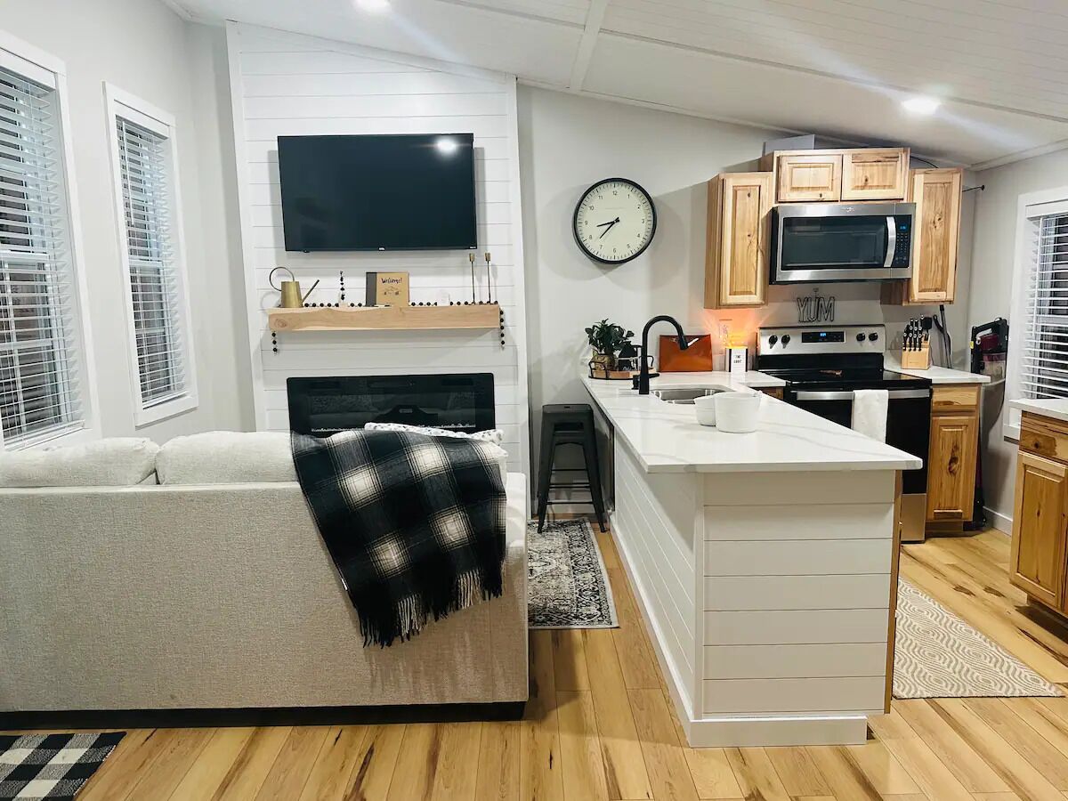 A modern living room with a sofa, a flat-screen TV, a fireplace, and a kitchen area with wooden cabinets and a microwave.