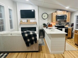 Una sala de estar moderna con un sofá, un televisor de pantalla plana, una chimenea y una zona de cocina con armarios de madera y un microondas.