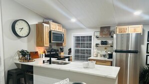 A modern kitchen with a central island, wooden cabinets, a stainless steel refrigerator, and a microwave. - Sunset Tiny House Retreat | Fire Pit & Starry Sky (Logan)