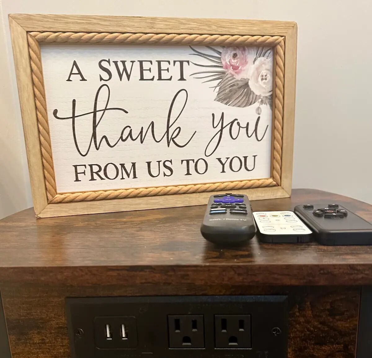 A framed sign with a message, a remote control, and a smartphone on a wooden surface with electrical outlets.