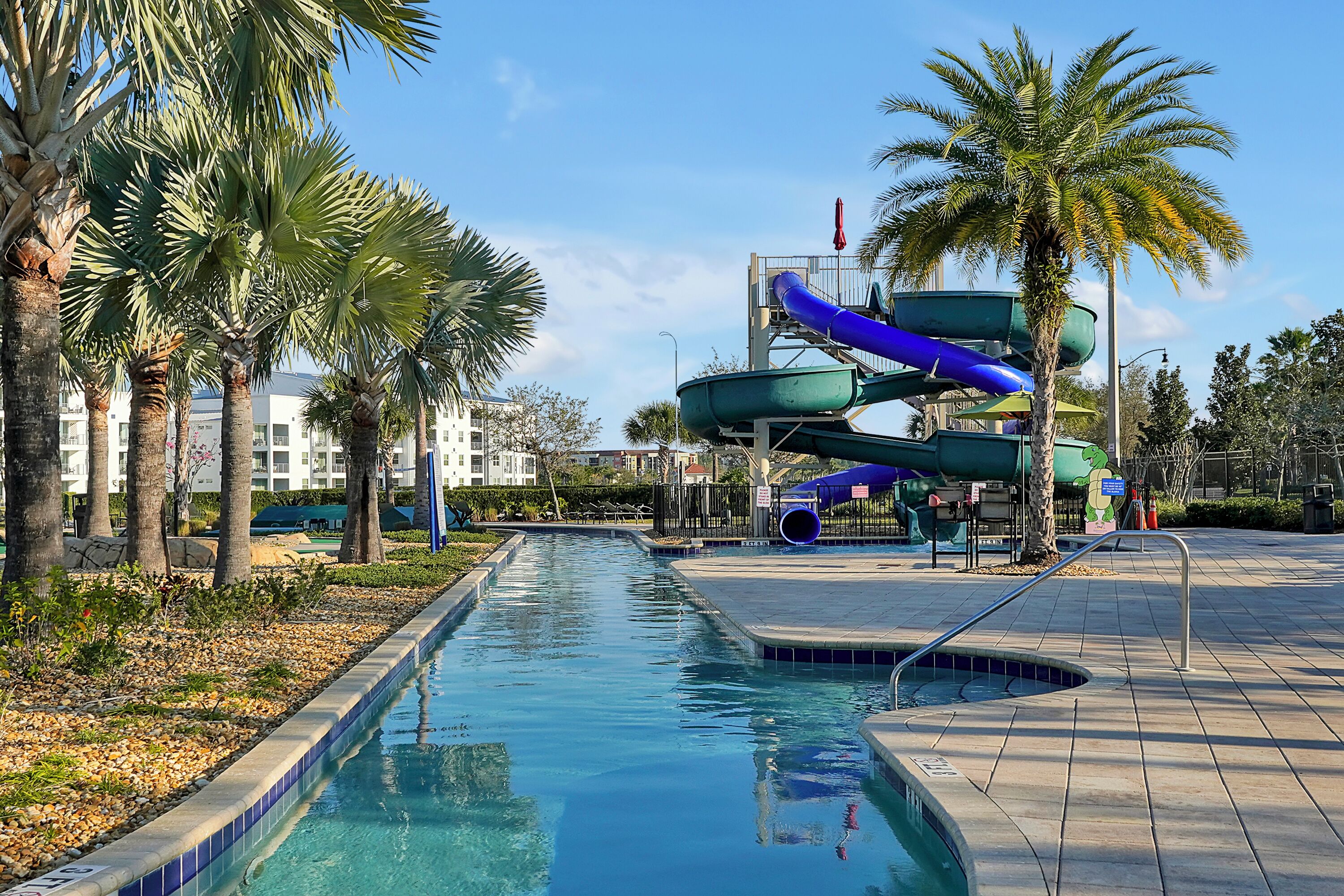 A swimming pool with a water slide, palm trees, and a building in the background.