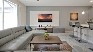 Ein modernes Wohnzimmer mit einer Eckcouch, einem Couchtisch mit Pflanze, einem an der Wand montierten Fernseher und einer Bar mit Hockern.