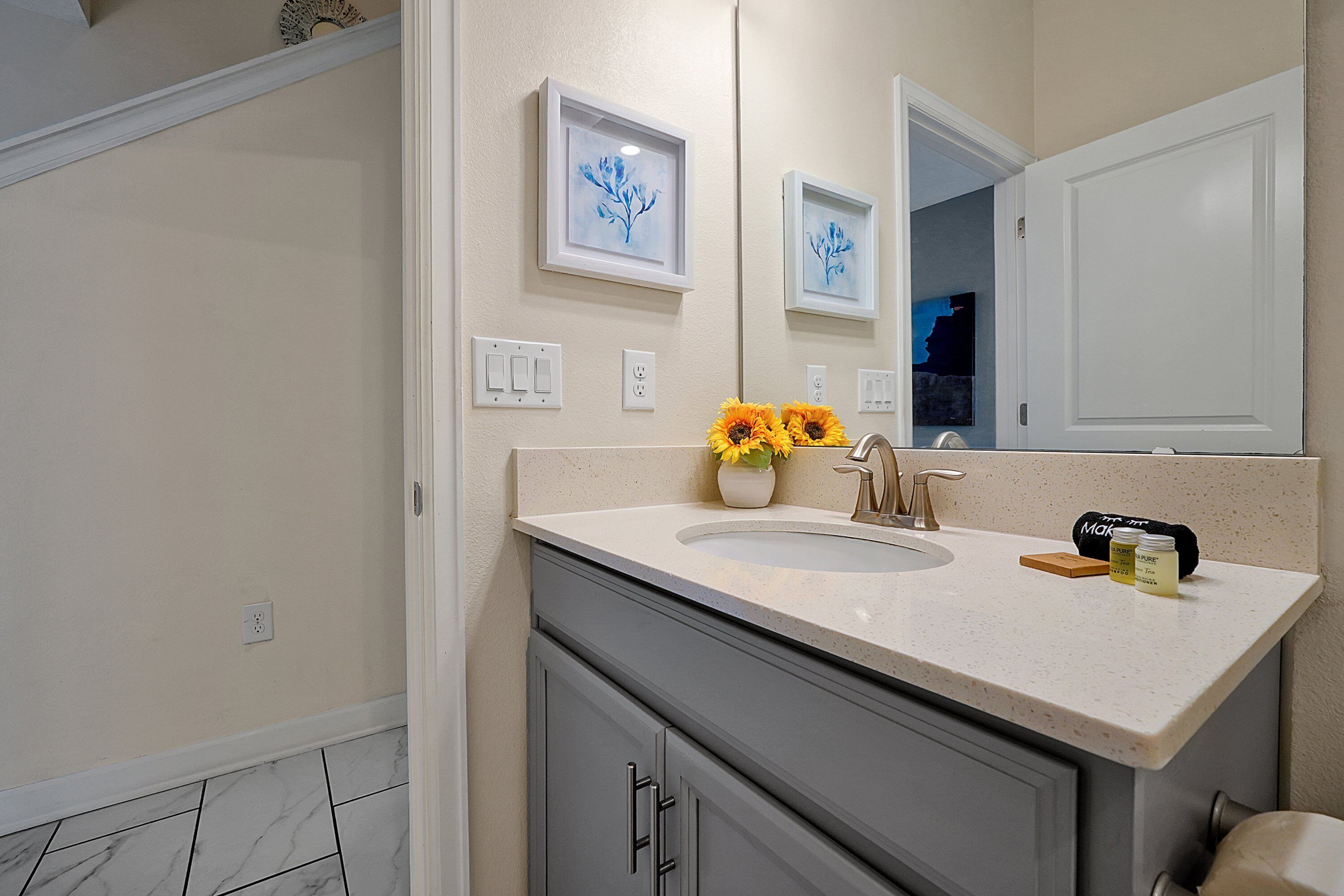 Une salle de bain avec un lavabo, un miroir, un tableau encadré et un vase de fleurs.