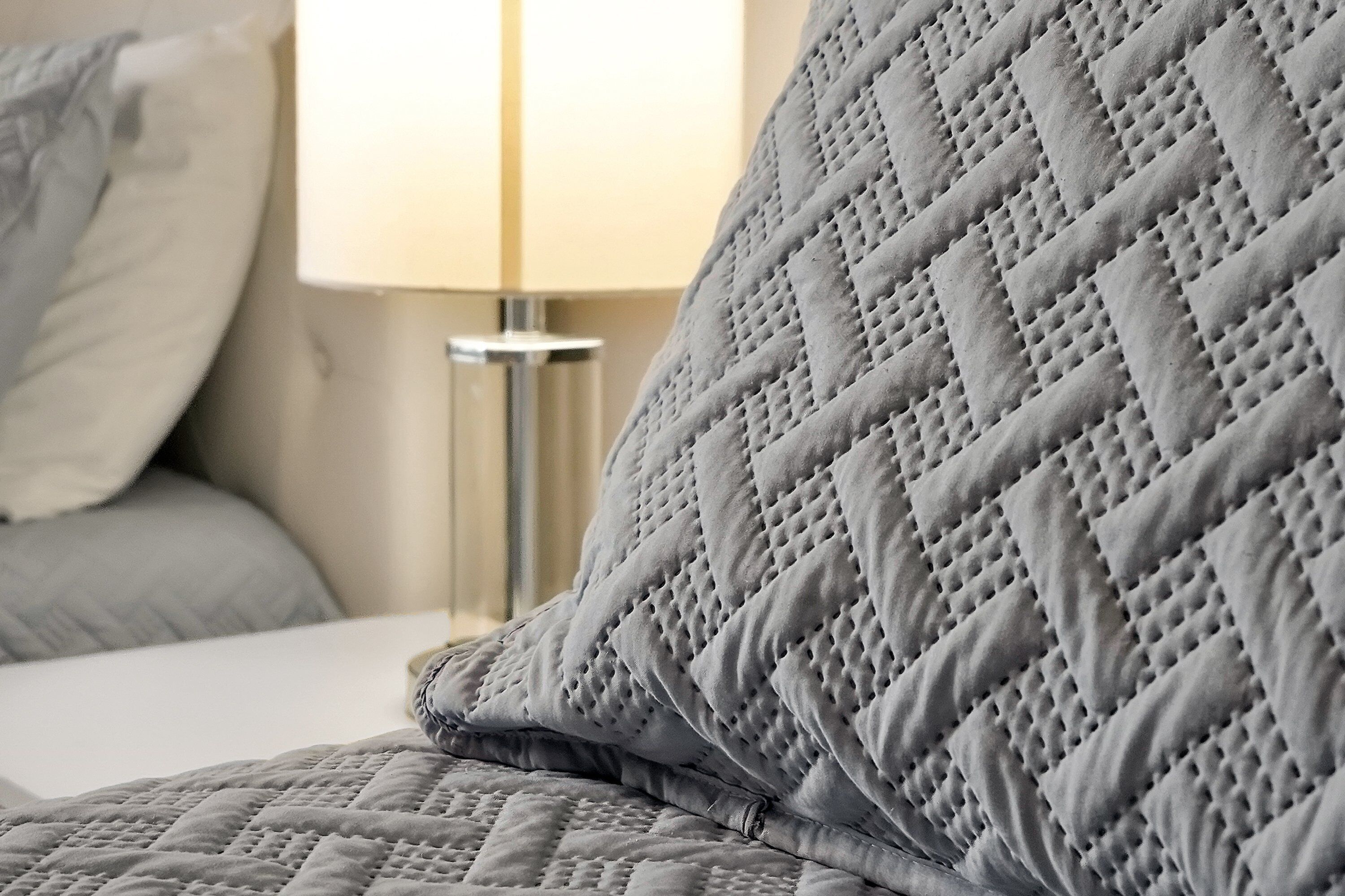 A bed with a quilted grey duvet and pillows, a bedside table with a lamp, and a window with curtains.