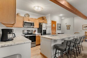 A modern kitchen with wooden cabinets, stainless steel appliances, a marble countertop, and a bar area with stools. - Across From Ski Lift, Hiking. Walk to Town. (Breckenridge)