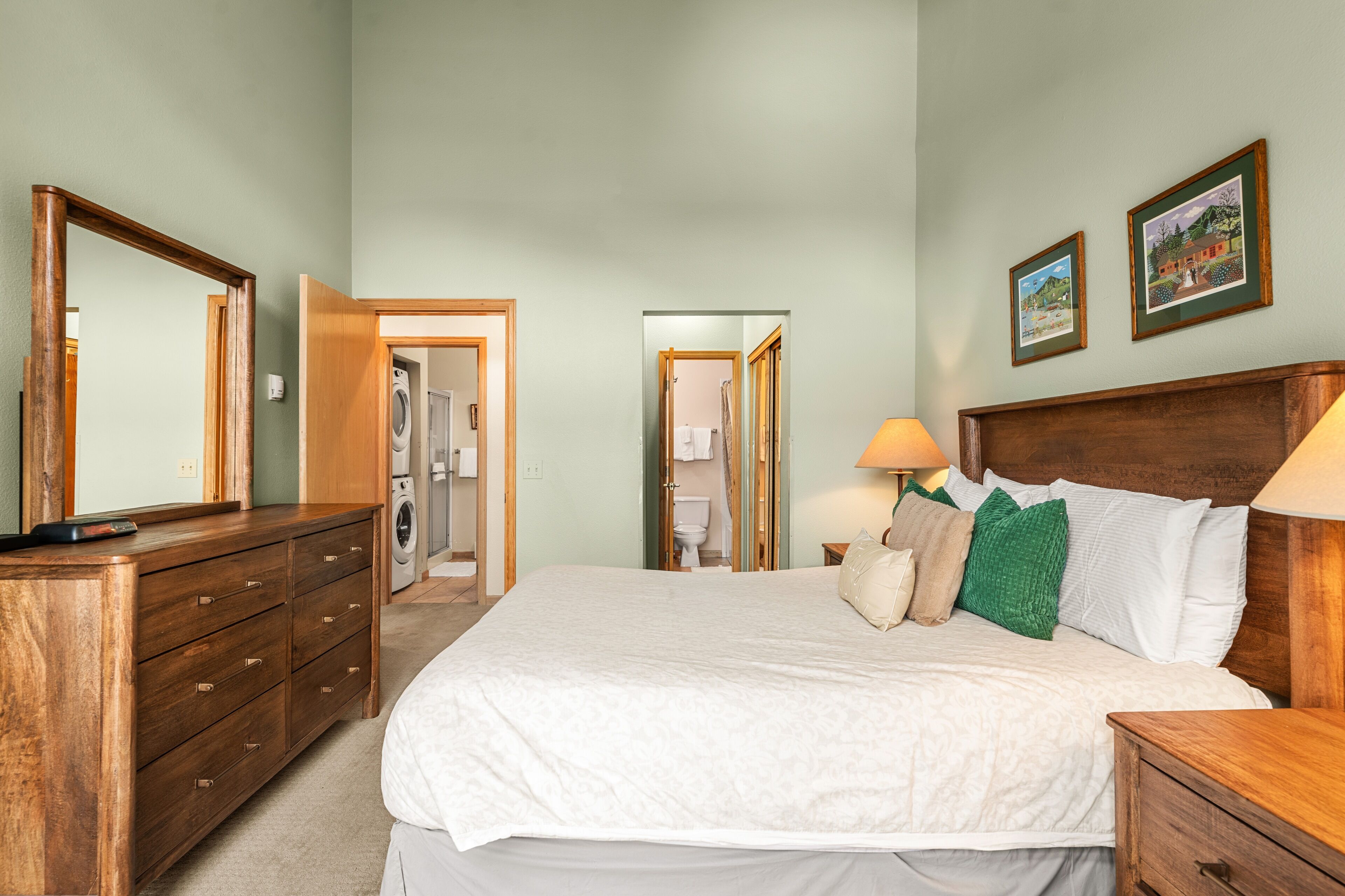 A bedroom with a large bed, wooden dresser, and a mirror. There is a door leading to a bathroom and a hallway.