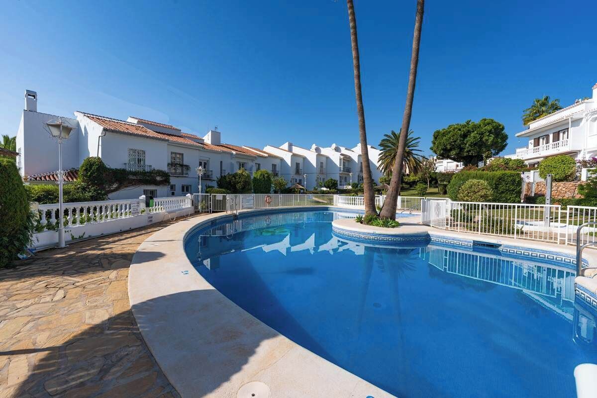 Une piscine entourée d’une terrasse en pierre, bordée de bâtiments blancs aux toits recouverts de tuiles rouges, de palmiers et surplombée d’un ciel bleu azur.