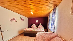 A bedroom with a wooden ceiling, a bed with floral bedding, a small bedside table, and a wall-mounted shelf.