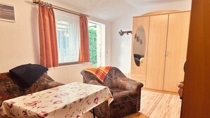 A cozy room with a wooden floor, a floral tablecloth-covered table, two patterned sofas, a wardrobe, and a window with curtains.