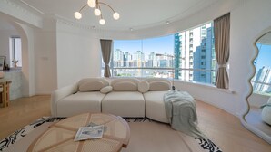 Ein modernes Wohnzimmer mit einer großen Couch, einem runden Couchtisch und Blick auf eine Stadtlandschaft durch hohe Fenster.