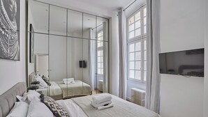 Quarto de hotel moderno com uma cama grande, um guarda-roupa com espelhos, uma televisão e vista para o exterior através de amplas janelas.