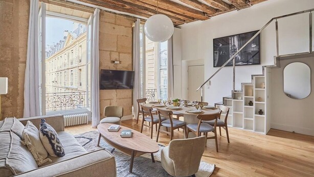 Uma sala de estar moderna com sofá, mesa de jantar e cadeiras, janelas amplas com vista para prédios, e uma escada com corrimão branco.