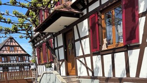 A traditional half-timbered building with a wooden door and red shutters, outdoor seating with a yellow table and plastic chairs, and a stone staircase leading to the entrance.