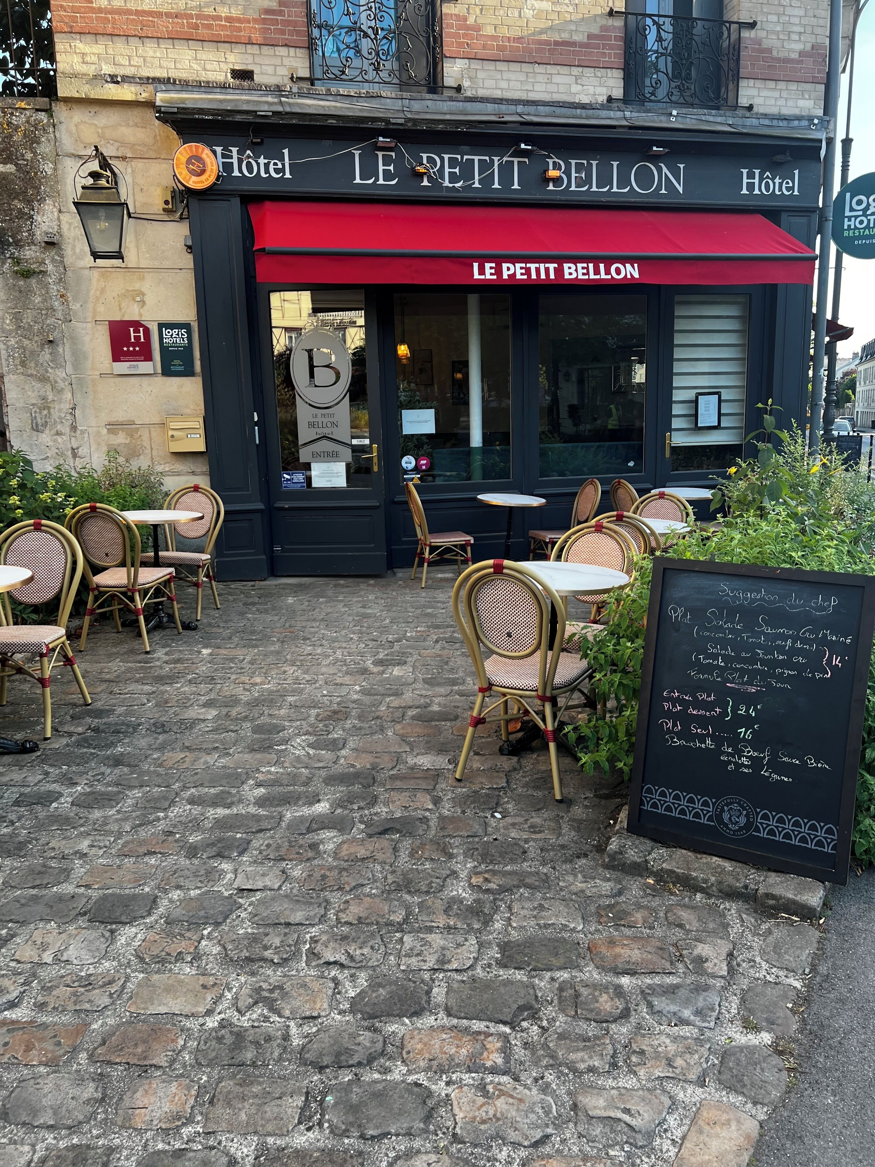 A hotel entrance with outdoor seating, a chalkboard menu, and a sign reading 'LE PETIT BELTON HOTEL.'