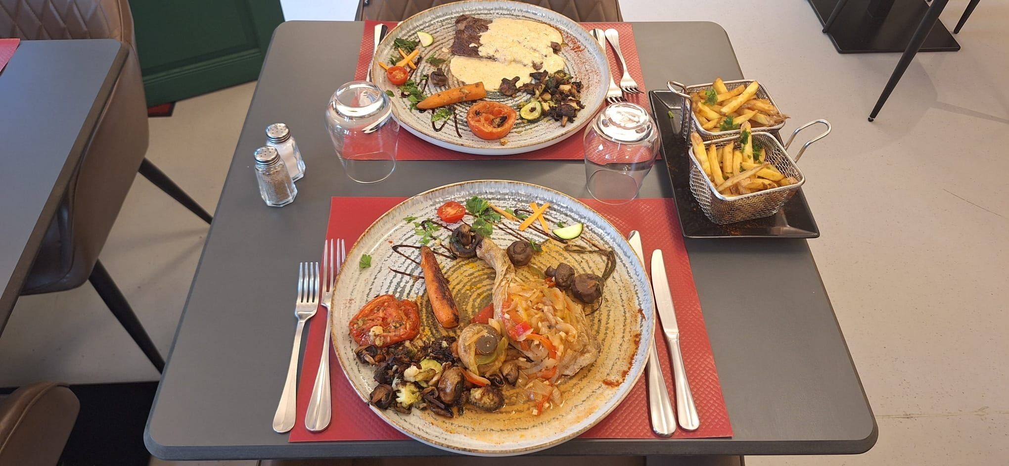 A dining table with two plates of food, salt and pepper shakers, a glass of water, and a basket of fries.