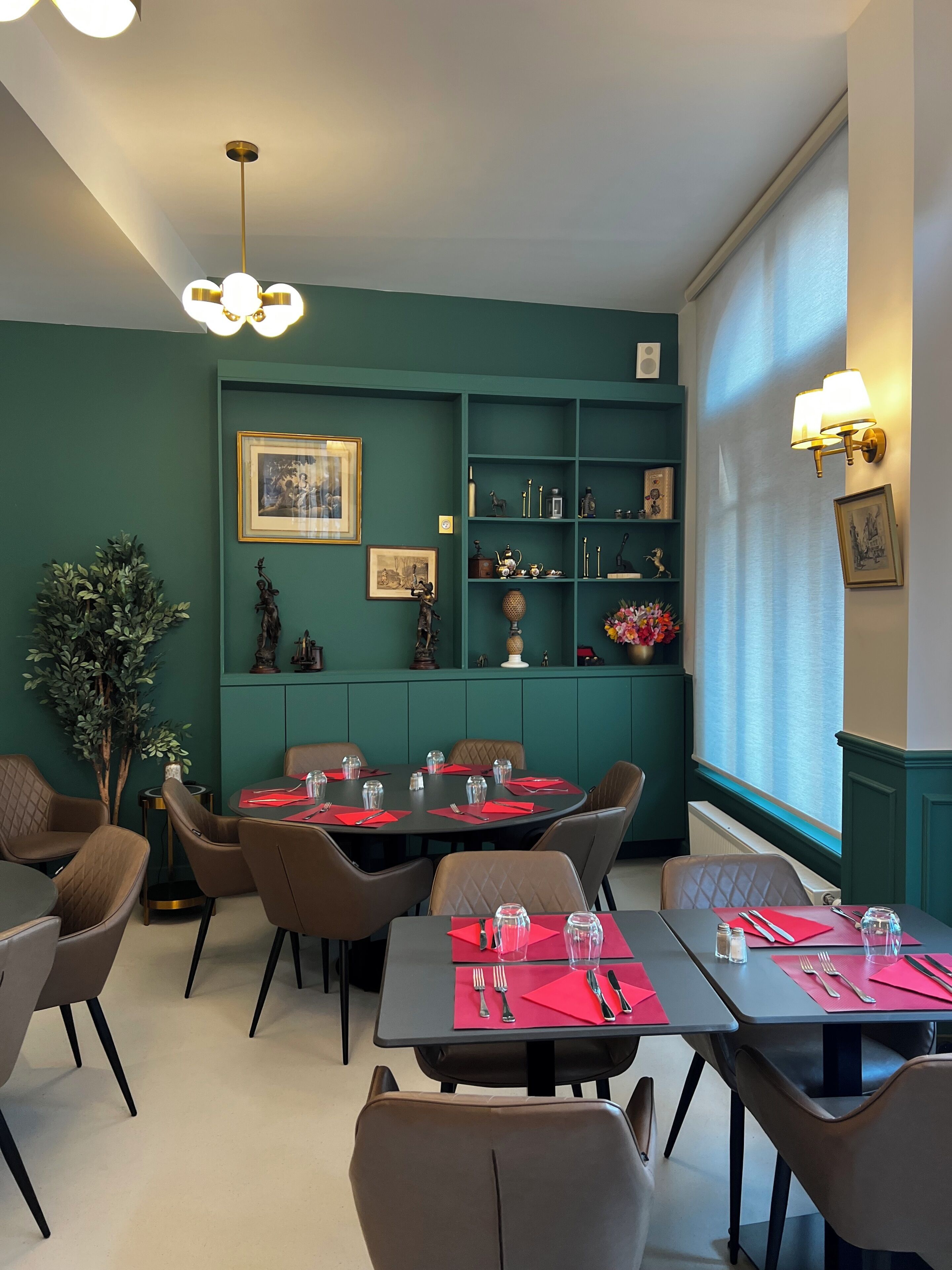 Comedor con paredes color turquesa, una araña y un ventanal con cortinas. Las mesas están dispuestas con servilletas y cubiertos rojos.