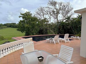 Un espace aménagé au bord de la piscine, avec des chaises longues blanches et une table en verre, entour é d’une terrasse carrelée et offrant une vue sur un paysage verdoyant luxuriant.