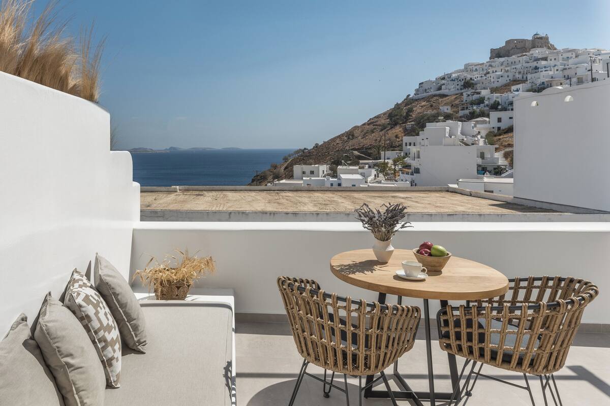 A terrace with a round table, wicker chairs, and a view of a coastal town with white buildings and a hill in the background.