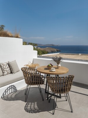 A terrace with a round table and four wicker chairs, a white sofa with patterned cushions, and a view of the sea and hills.