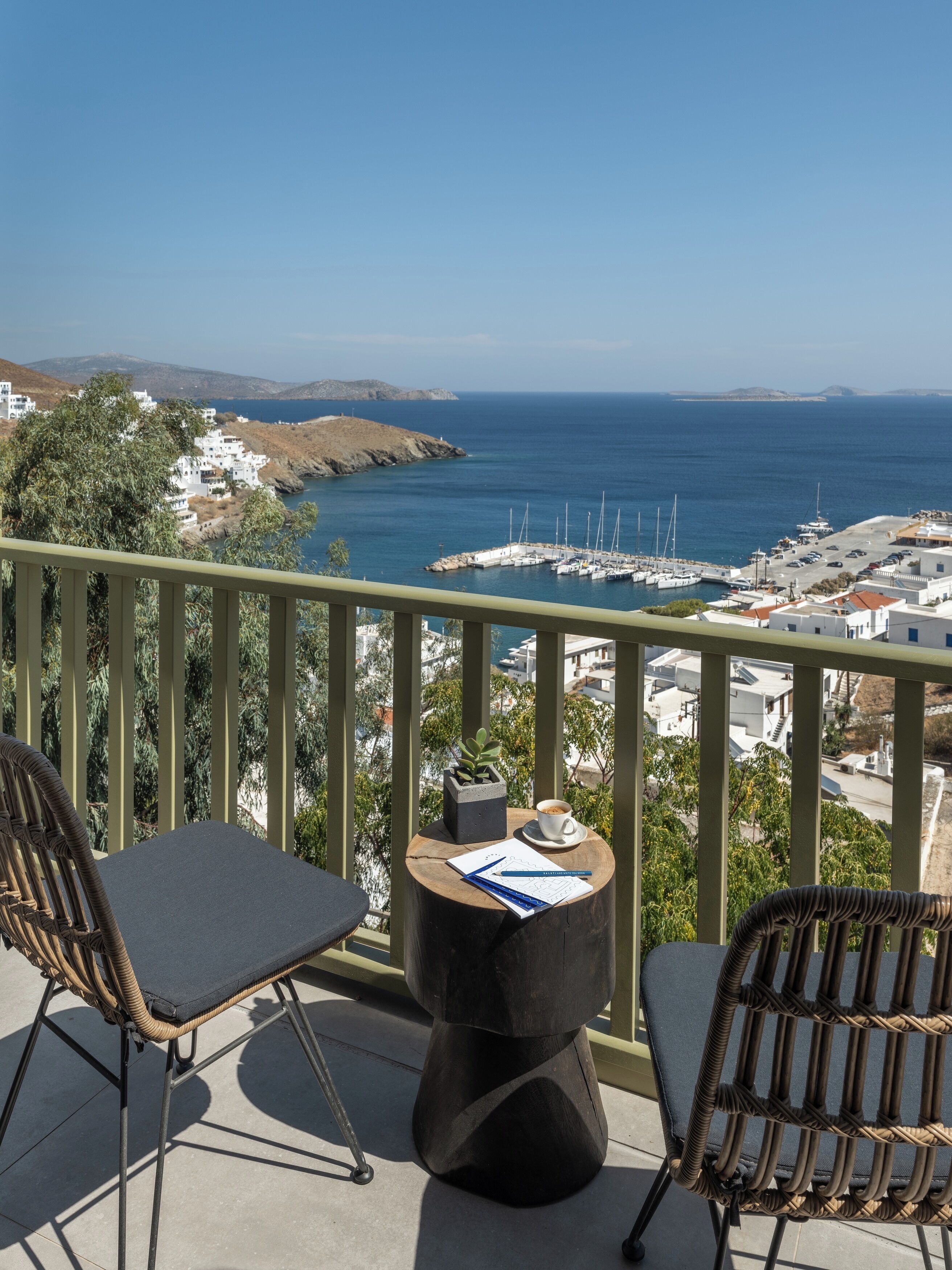 Ein Balkon mit Korbstühlen, einem kleinen Tisch mit Tasse und Papieren, und Blick auf eine Küstenstadt mit weißen Häusern und einem Hafen.