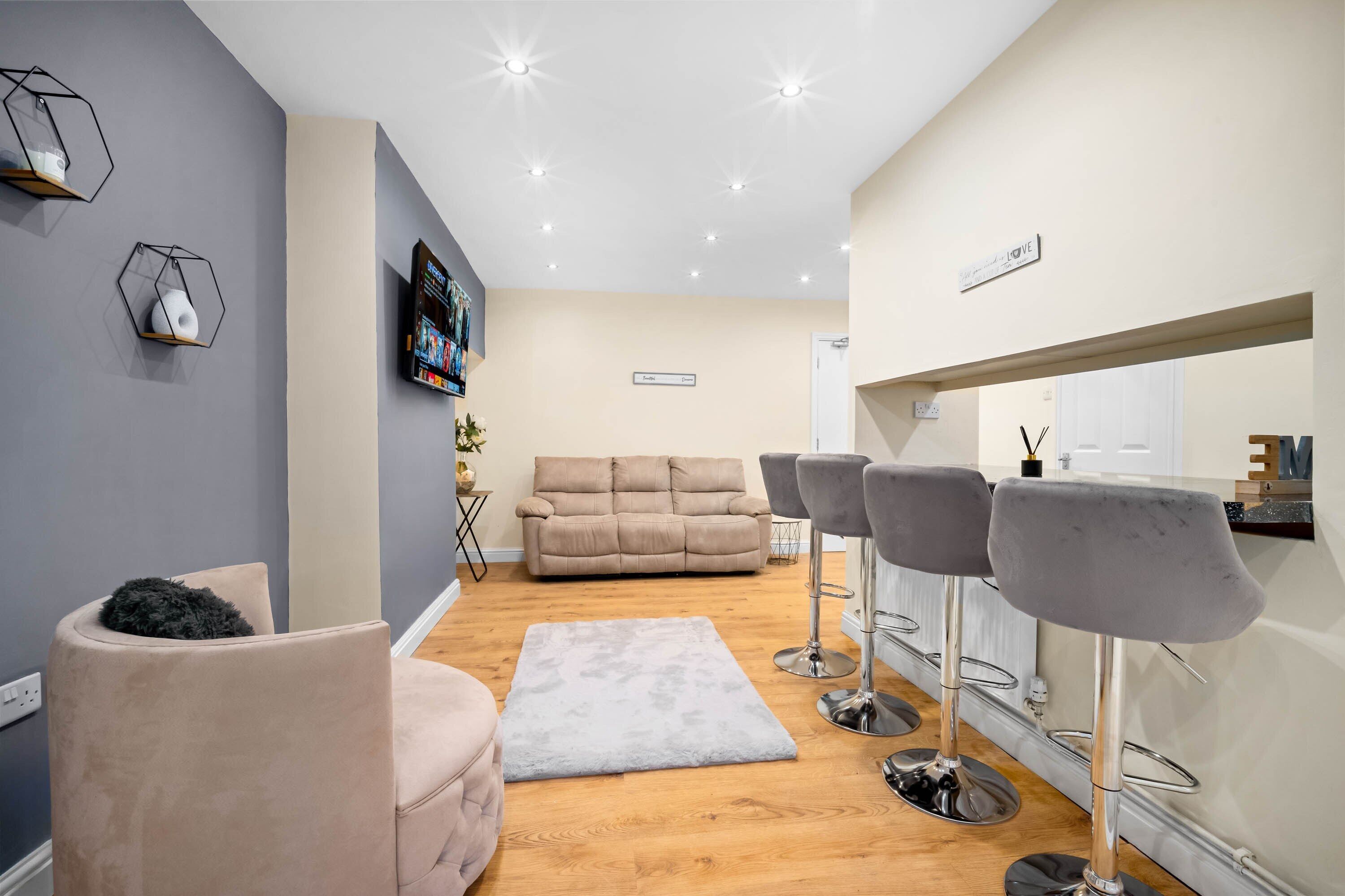 A modern living room with a sofa, a coffee table, and a TV mounted on the wall. There are two bar stools and a small kitchen area in the background.
