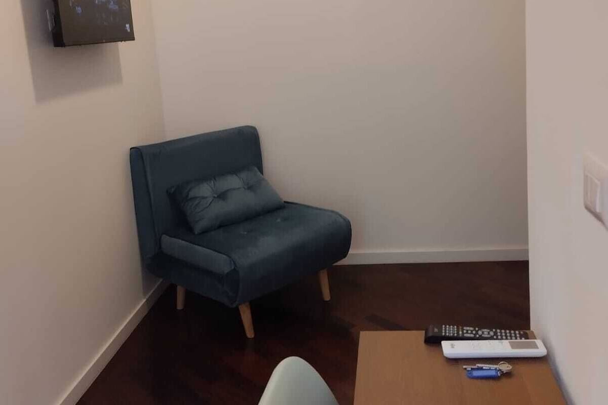 A small room with a blue armchair, a wooden table, and a TV mounted on the wall.
