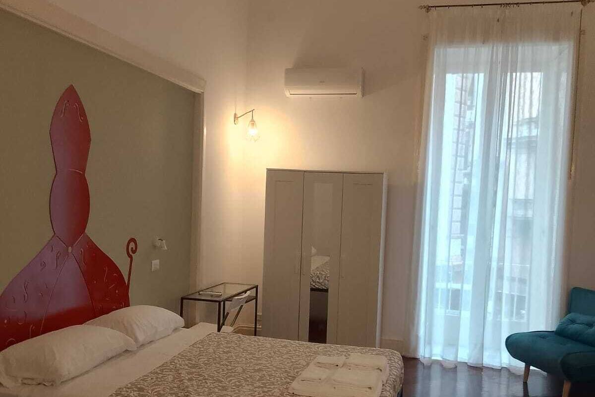 A bedroom with a bed, a wardrobe, a small table, a chair, and a window with curtains.