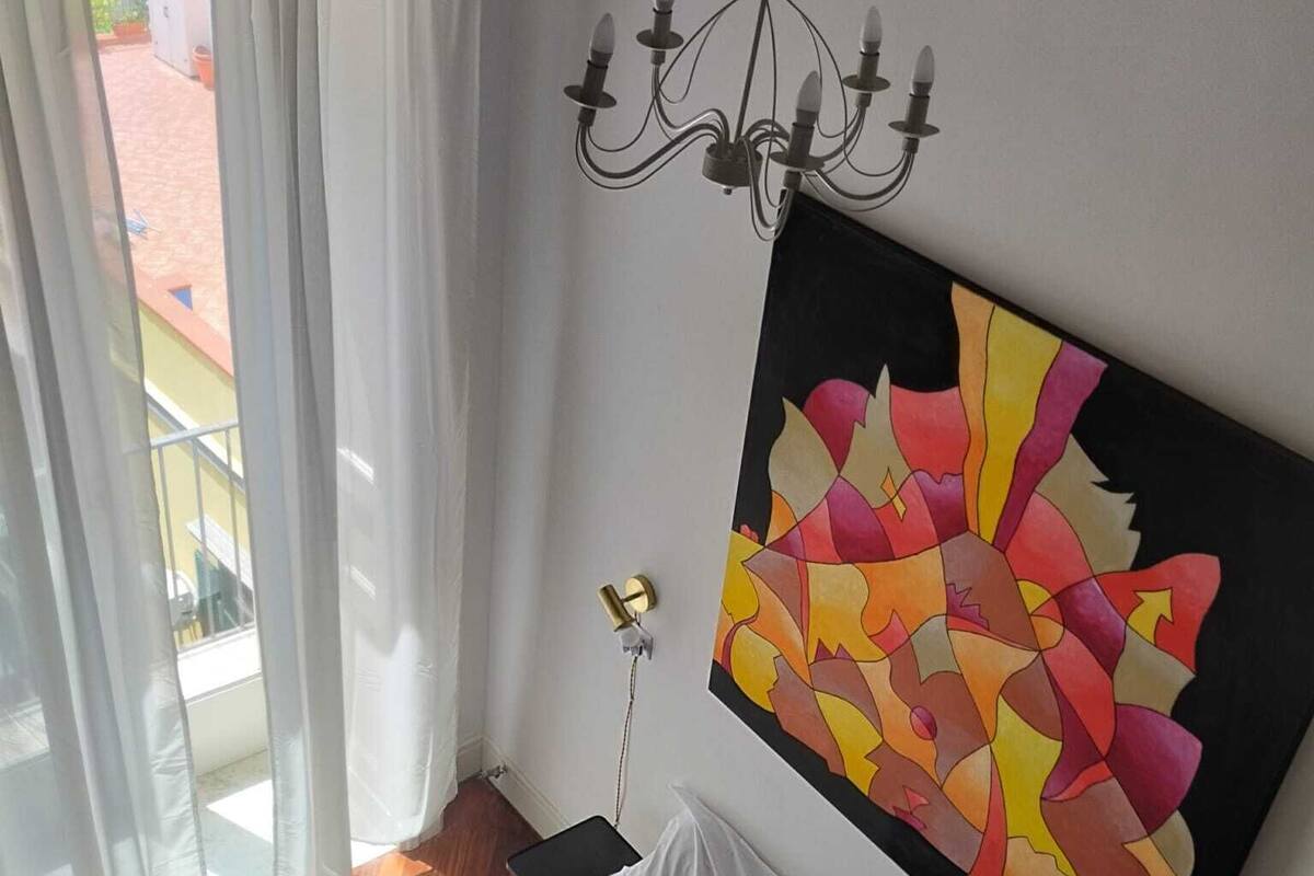 A bedroom with a bed, a chandelier, a large abstract painting, and a view through a window with curtains.