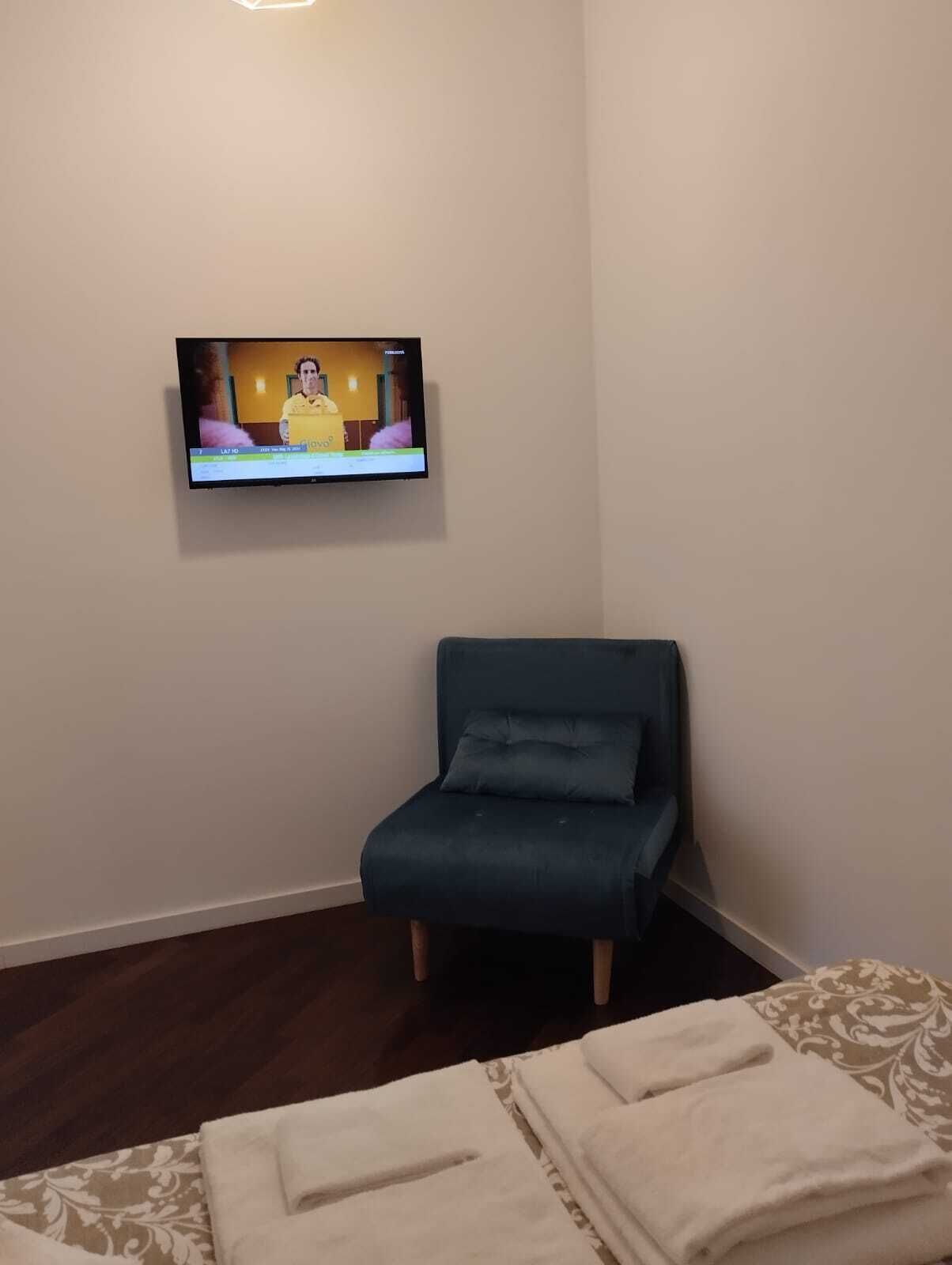 Una habitación con un televisor de pantalla plana montado en la pared, un sillón azul oscuro y una cama con colcha estampada.
