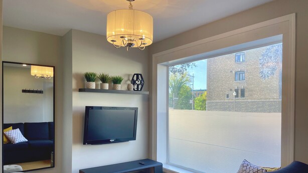 A modern living room with a large TV, a dark sofa, a small ottoman, a console table, and a large window with a view of a building. - 29SV-VE Newo Retro (Quebec)