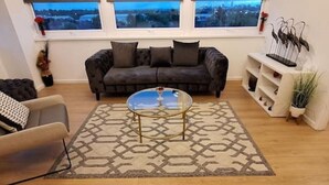 Una sala de estar con un sofá marrón oscuro, una mesa de centro de vidrio y una alfombra estampada.