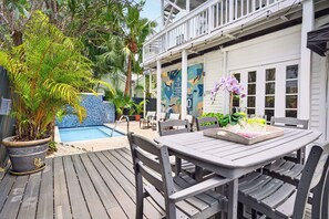 A wooden deck with a table and chairs, a potted plant, a pool, and a white building with a balcony.