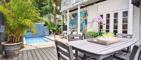 Um deck de madeira com mesa e cadeiras, um vaso de planta, uma piscina e um edifício branco com varanda.