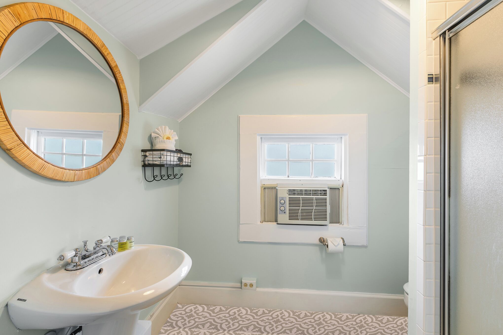 Une salle de bain avec un lavabo blanc, un grand miroir, une fenêtre avec un appareil de climatisation et des carreaux de sol à motifs.