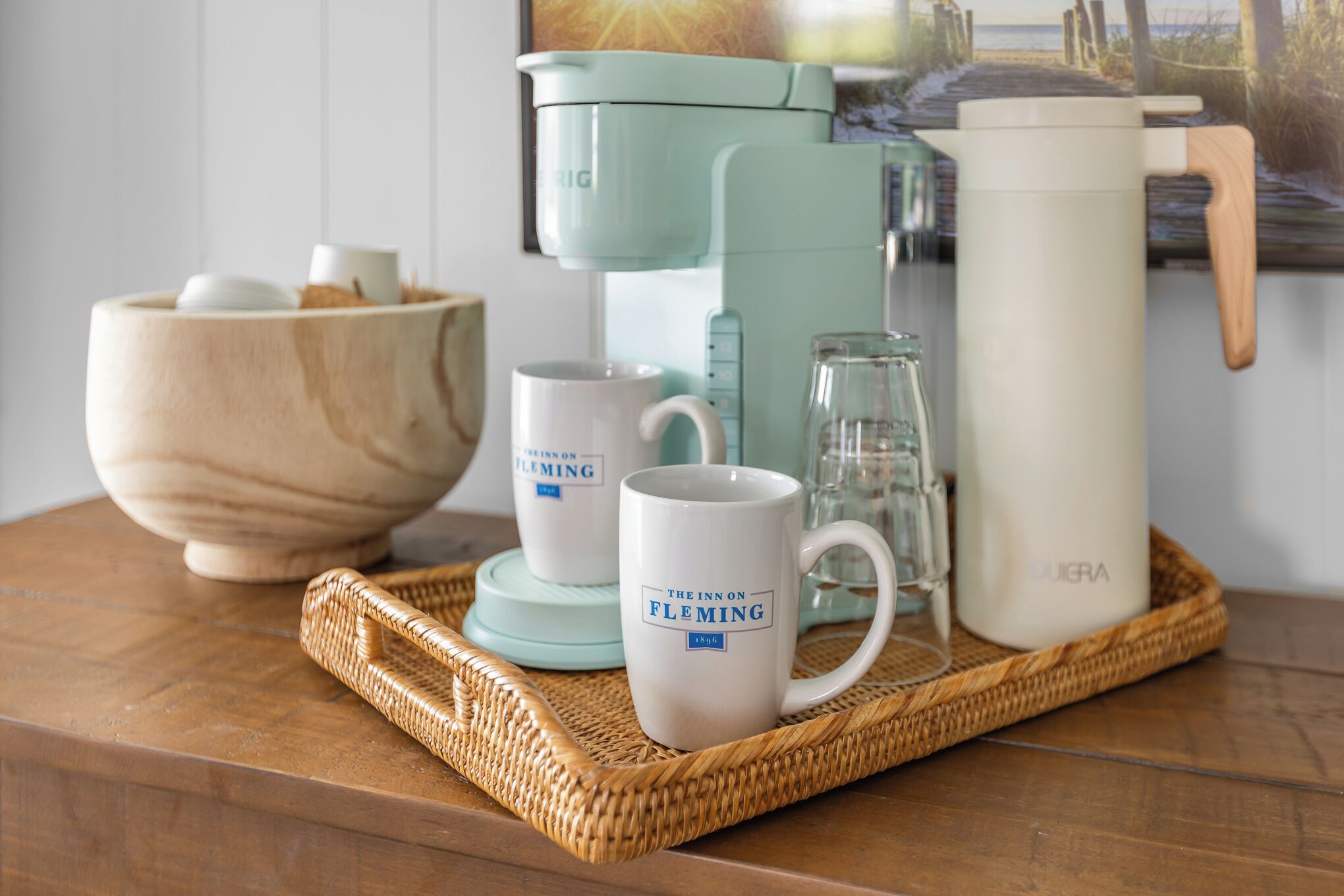 Una encimera de cocina con una cafetera, dos tazas, un vaso, un bol de madera y una jarra blanca.