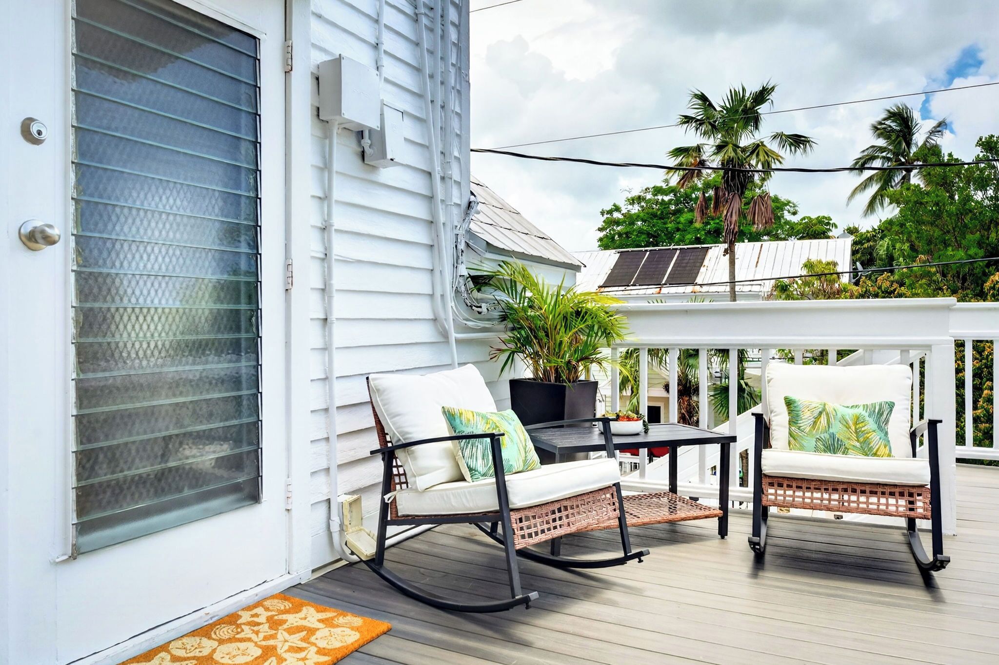 A covered outdoor area with a white building, two rocking chairs, a table, and a potted plant.