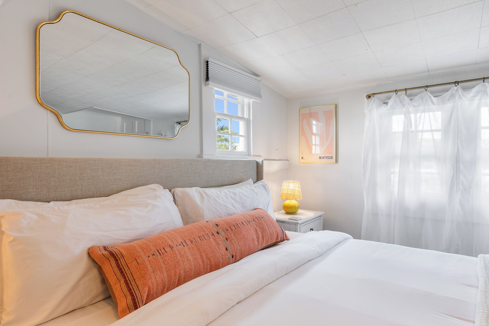 Een net opgemaakt bed met witte en oranje kussens, een bedlampje en een grote spiegel aan de muur.