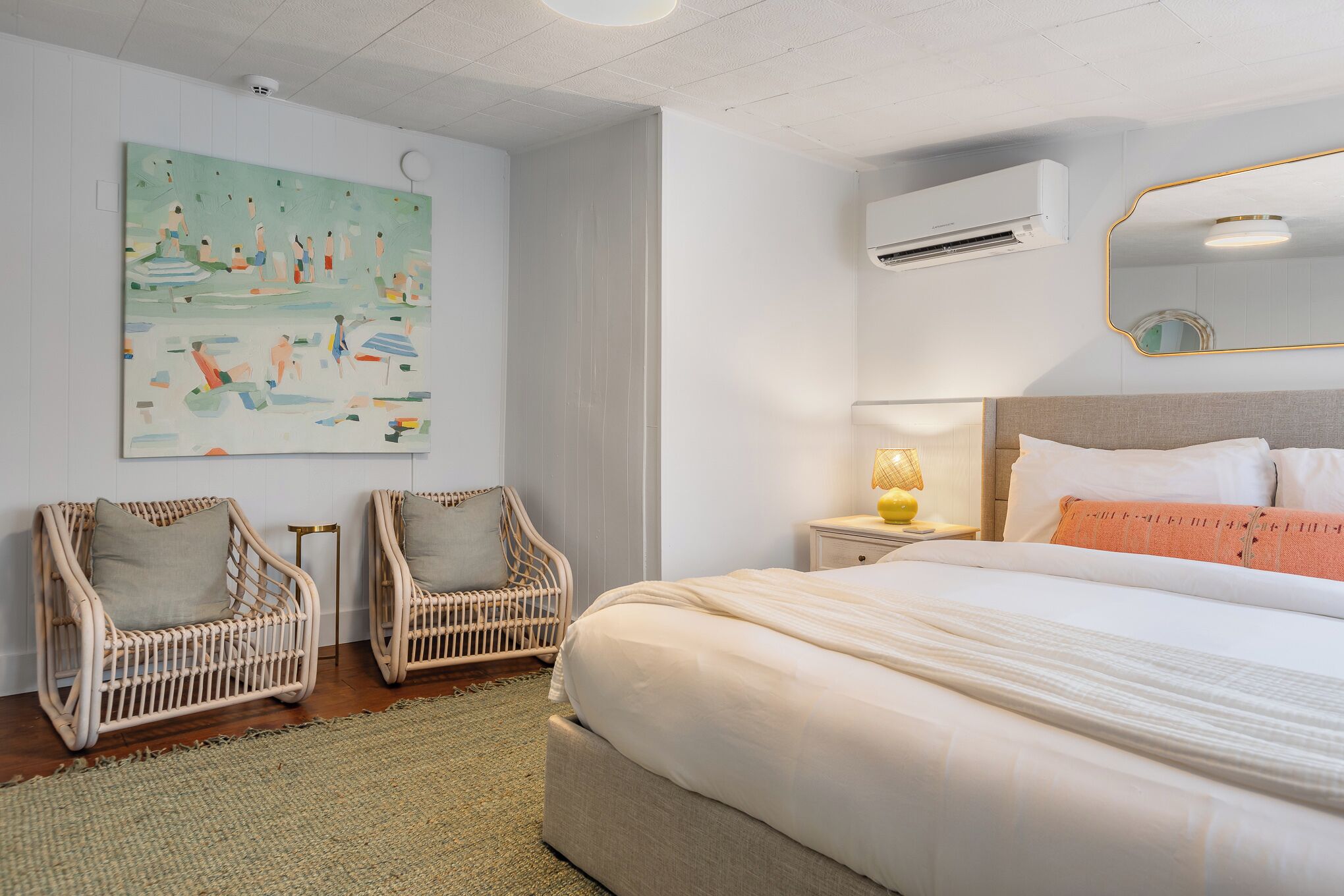 A bedroom with a large bed, two wicker chairs, a painting on the wall, an air conditioning unit, and a bedside lamp.