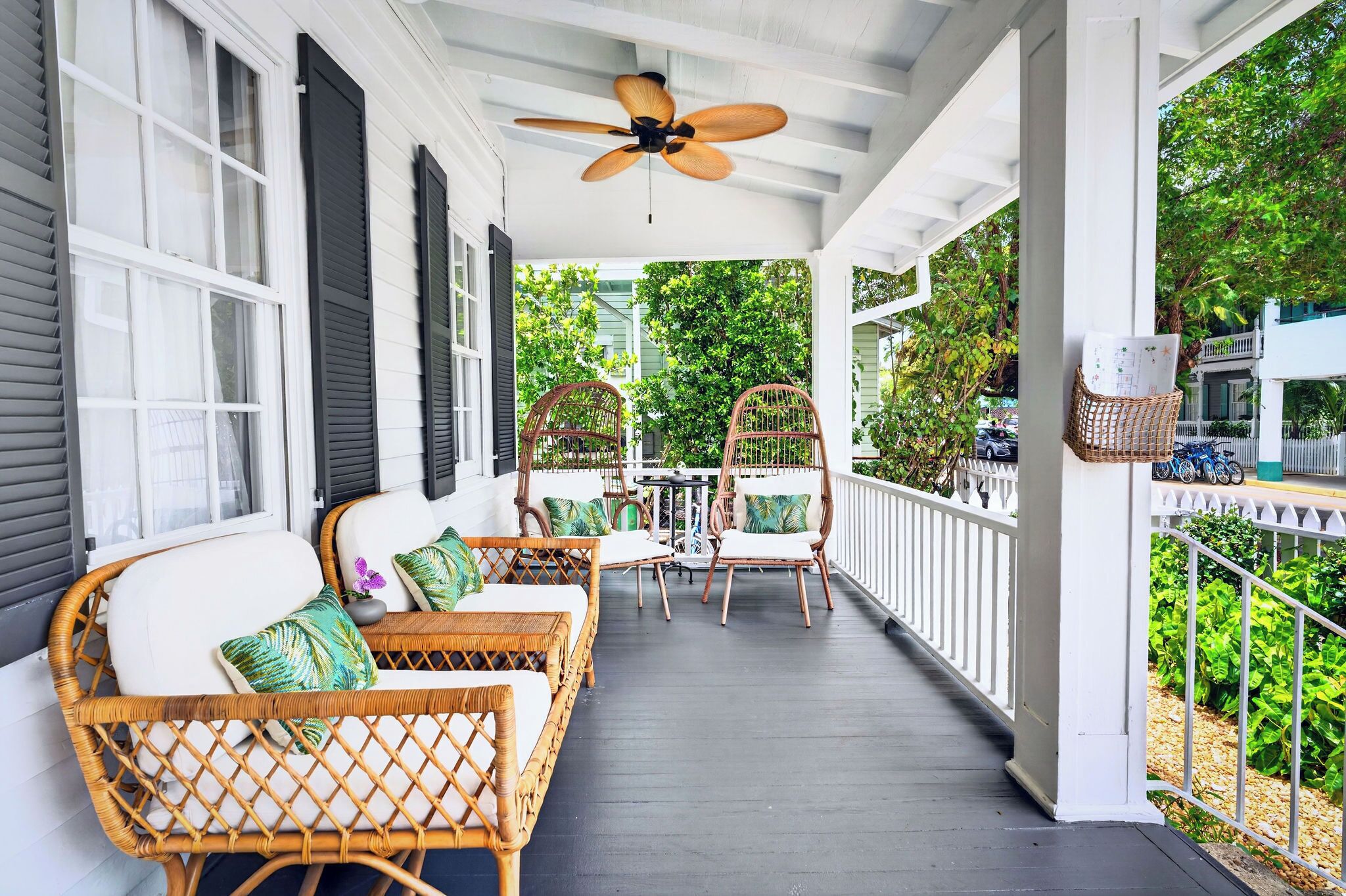 A covered porch with wicker furniture, a ceiling fan, and a view of greenery.