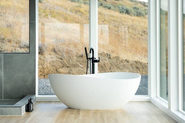 A modern bathroom with a freestanding bathtub, large windows, and a view of a natural landscape.