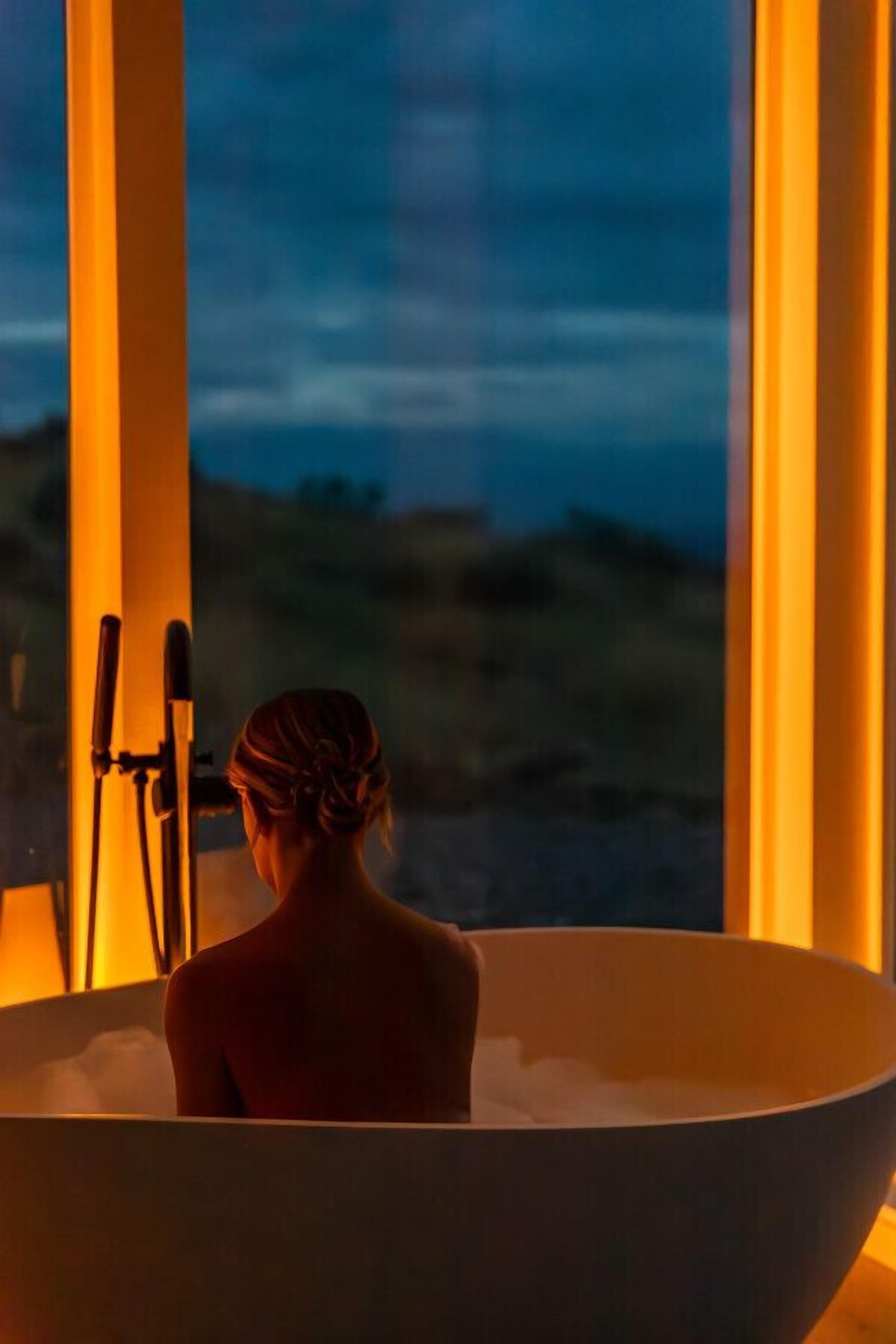 Une personne dans une grande baignoire, avec vue sur un paysage à travers de grandes fenêtres.