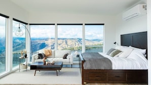 A modern bedroom with a large bed, a sofa, and a coffee table. There are large windows offering a view of the mountains.