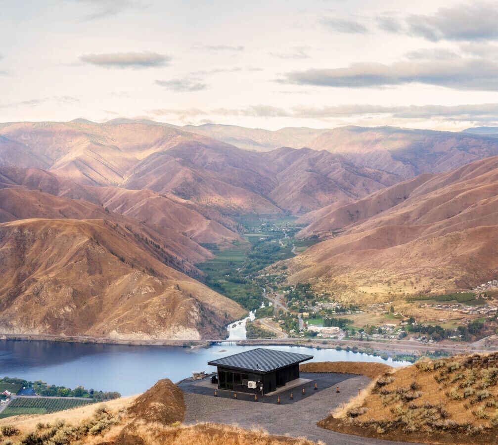 Un paysage montagneux avec un petit bâtiment situé près d’une étendue d’eau.
