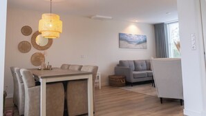 A modern living room with a dining area, a grey sofa, and a large window with curtains. - Julia's Holidayhome (Egmond-Binnen)